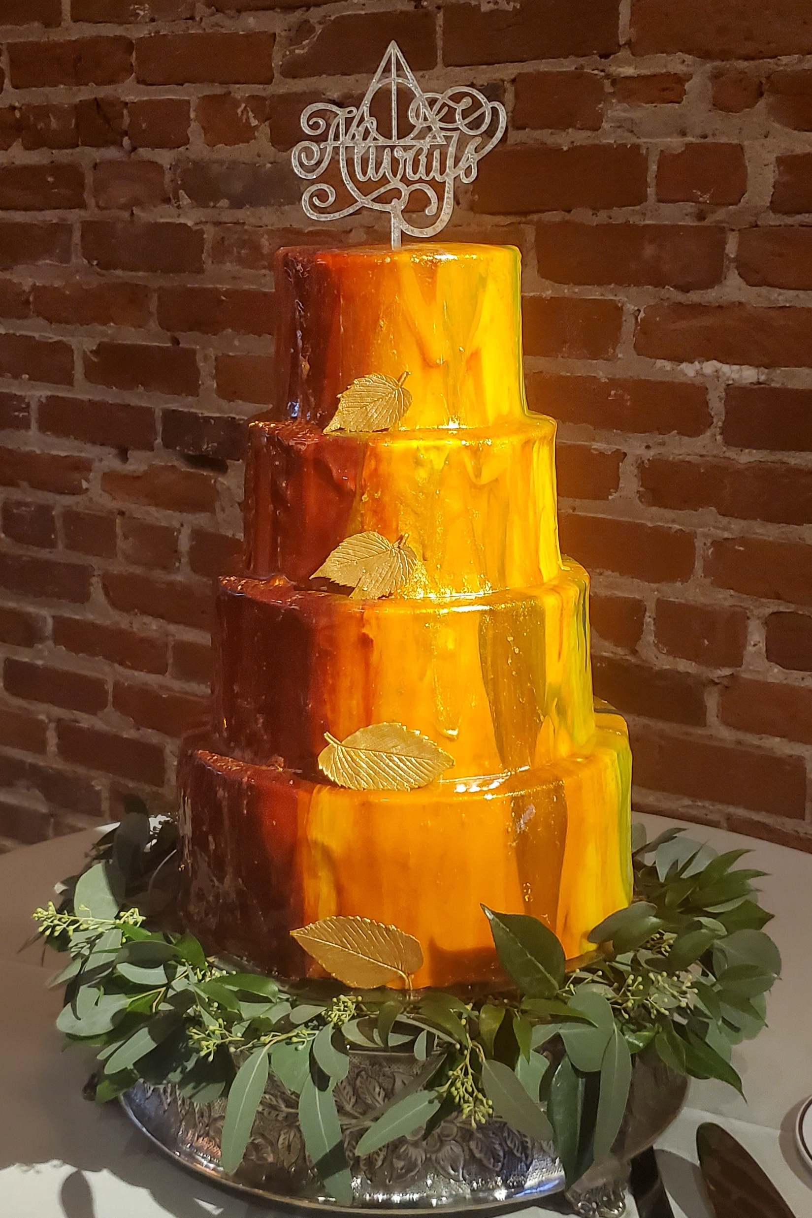 A four-tiered wedding cake in front of an exposed brick wall. The cake has been covered in mirror glacage in fall colors, and there is a golden chocolate leaf on each tier. The topper says "Always"